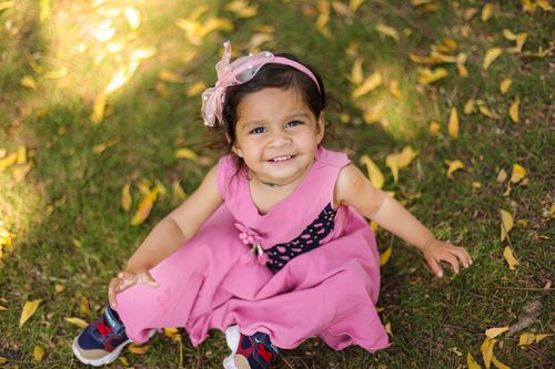 Free Charming young girl smiling joyfully while sitting on grass in a pink outfit with a bow. Stock Photo