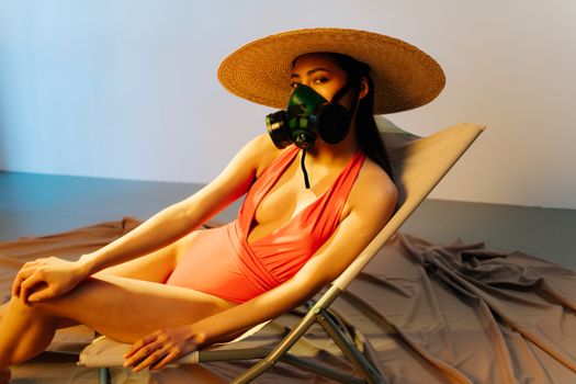 Stylish woman in swimsuit and gas mask reclines on a beach chair, blending fashion with a pollution concept.