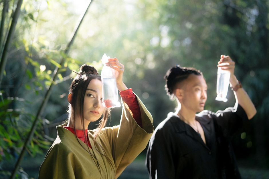 Two people in traditional attire hold bags with fish in a tranquil Japanese garden amidst lush greenery and sunlight.
