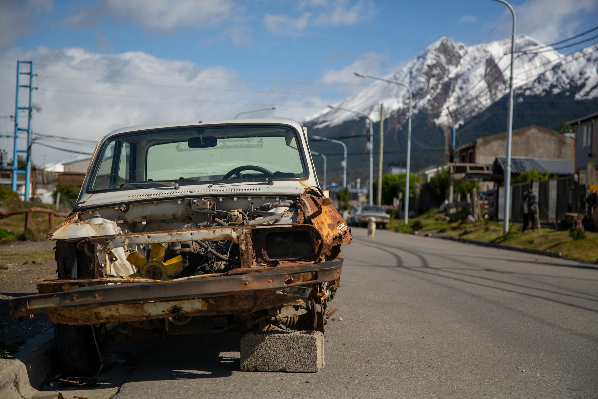 A rusty, broken car wreck abandoned on a quiet roadside with a mountain backdrop.