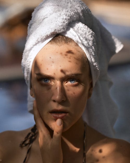 Young woman in terry towel on head with shadows on face touching chin while looking at camera in sun ray