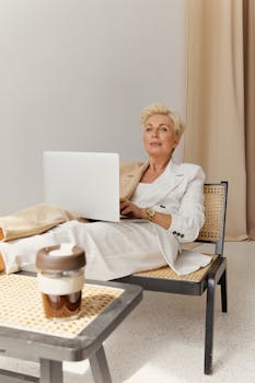A professional woman in a blazer relaxes while working from home on her laptop, sitting in a stylish indoor setting.