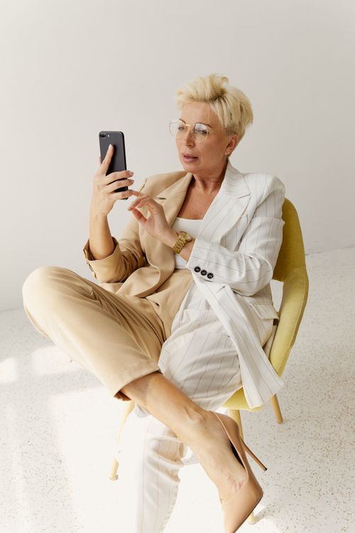 Free Elegant middle-aged businesswoman in a chic outfit sitting on a chair using a smartphone indoors. Stock Photo