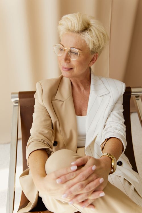 Free Elegant businesswoman with short blonde hair and eyeglasses sitting confidently in a chic setting. Stock Photo