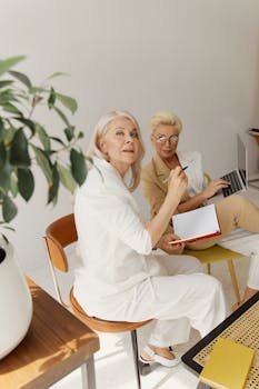 Two senior businesswomen collaborating with a laptop and notebook in a bright office setting.