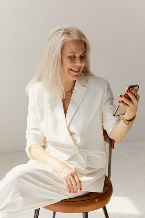 Free A stylish senior woman in white business attire using a smartphone indoors, smiling. Stock Photo