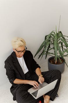 A trendy businesswoman in pinstripe suit works on laptop next to potted plant, embodying style and productivity.