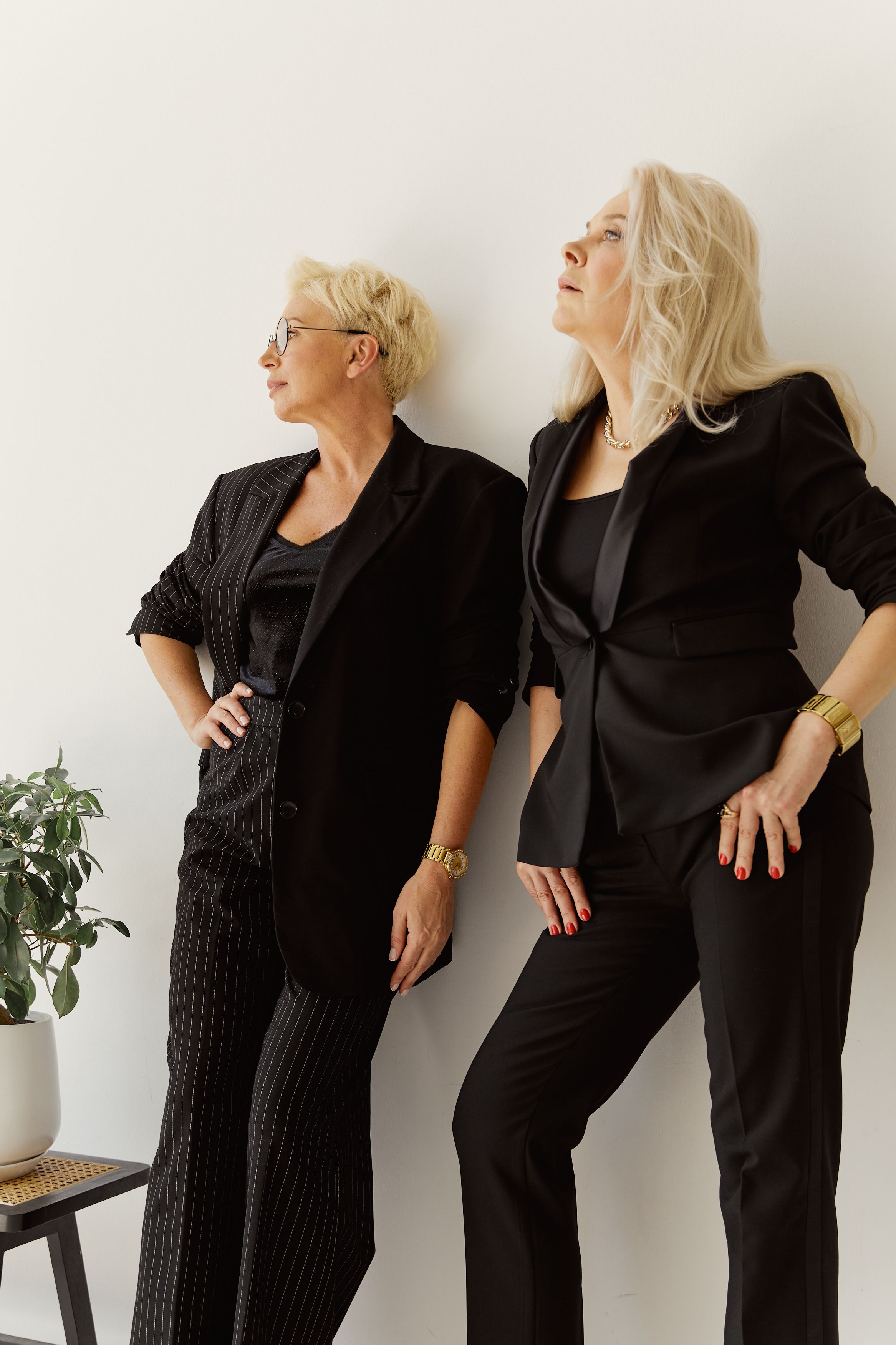 Two stylish women in black blazers and pants pose confidently indoors against a light background.