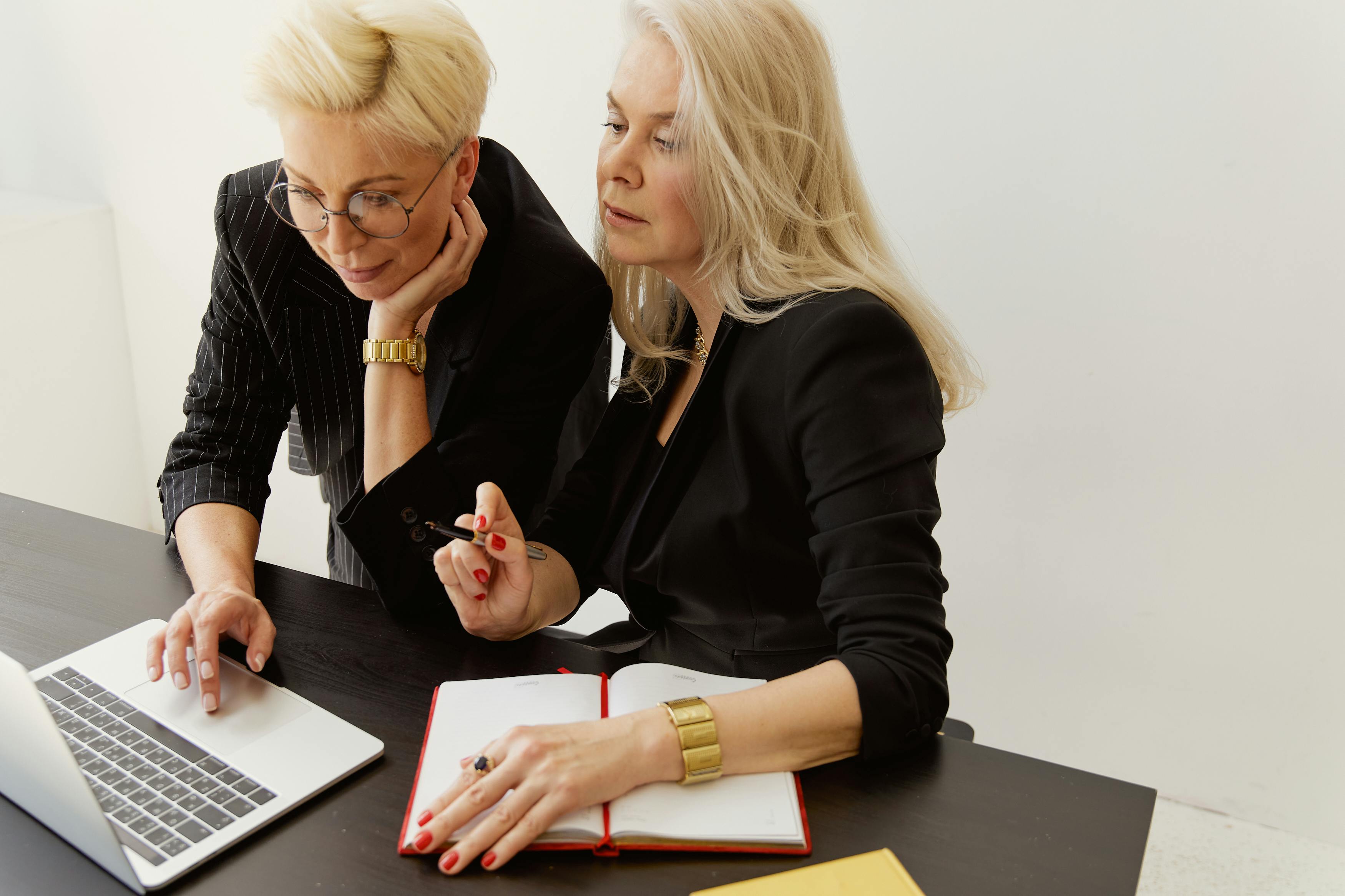 Two senior businesswomen working together at a laptop with notebooks and pens in hand.