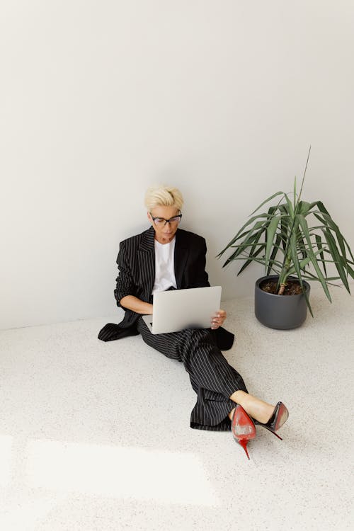 Free Stylish businesswoman in striped suit working on a laptop near a plant in a minimalistic indoor setting. Stock Photo