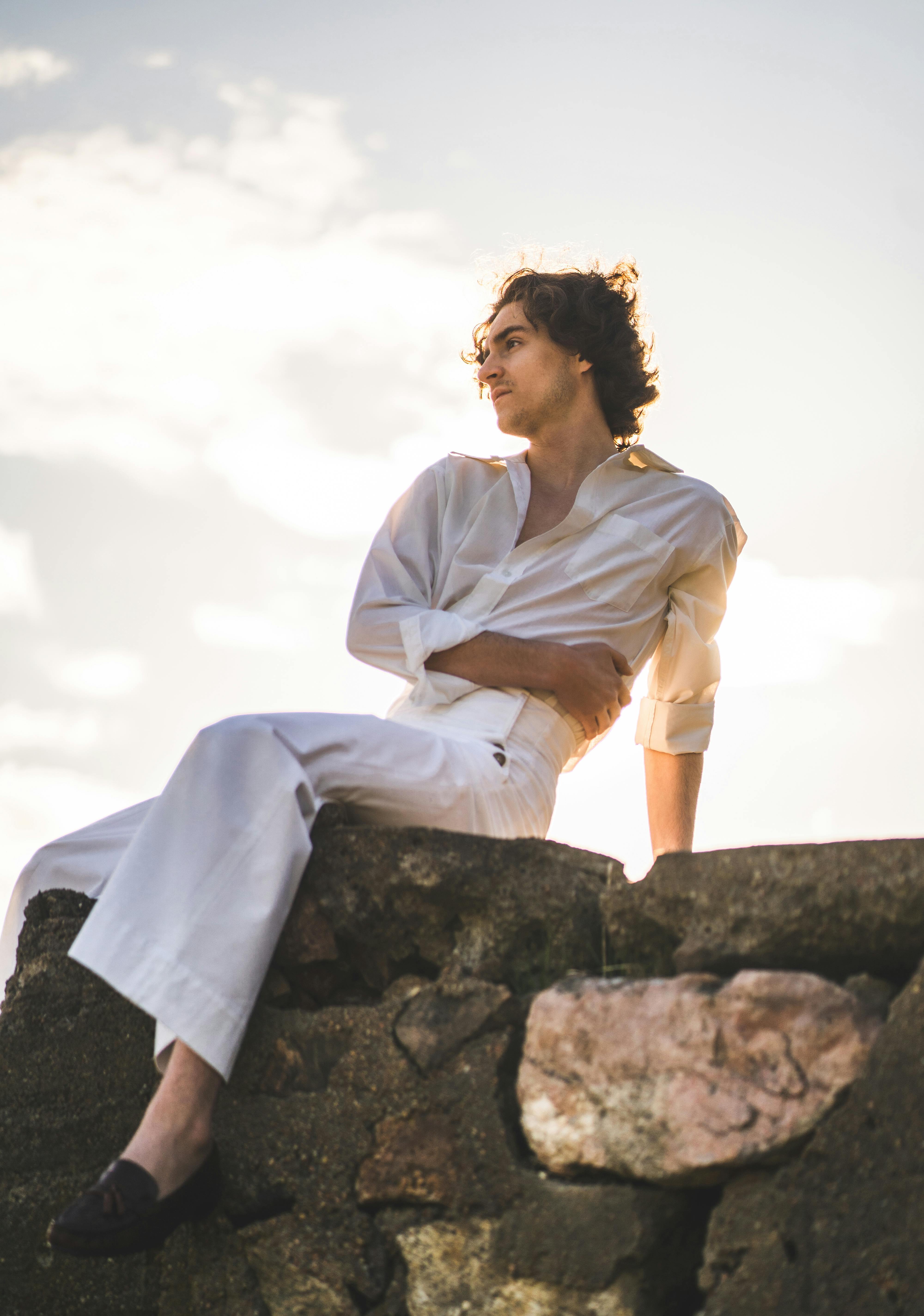 A man in a fashionable white outfit poses on a stone wall under a bright sky.
