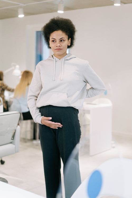 Free Pregnant woman standing in an office, hand on belly, wearing a hoodie. Modern workspace environment. Stock Photo