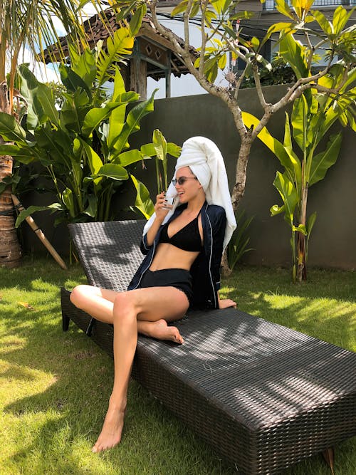 Free Full body of gorgeous young female in black clothes and towel on head resting on deckchair and drinking water in summer tropical garden Stock Photo