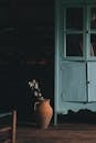 Old fashioned cupboard placed on wooden floor near white flowers in clay vase in dark room