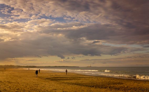 A tranquil beach scene at sunset with scattered clouds and a distant lighthouse, creating a calming atmosphere.