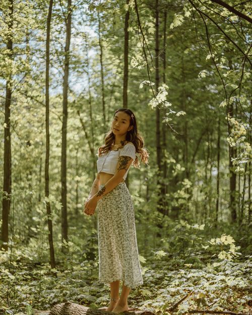 Free Full body of young ethnic female with tattoos standing on trunk among trees covered with green foliage in woods and looking away Stock Photo