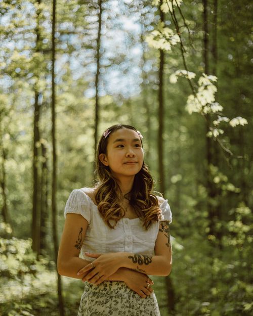 Free Positive charming Asian female with long wavy hair standing with crossed arms in woods and looking away Stock Photo