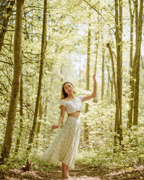 Free Full body of young barefooted ethnic female in long skirt and top standing with raised arm in sunny woodland Stock Photo
