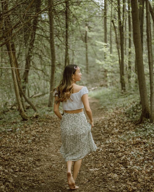 Back view of positive ethnic female in skirt strolling on path with fallen dry leaves in woodland