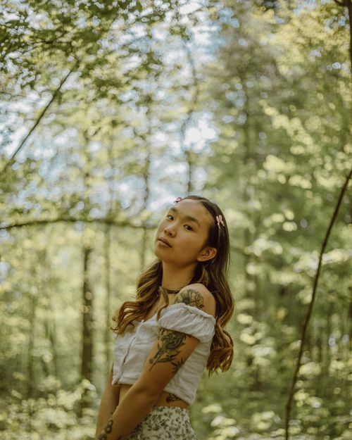 Free Side view of charming young ethnic female in top standing in forest among trees and looking at camera Stock Photo