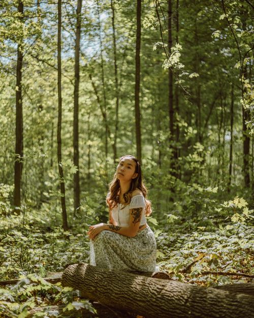 Free Side view of ethnic female with tattoos sitting on fallen trunk among green plants and looking at camera Stock Photo
