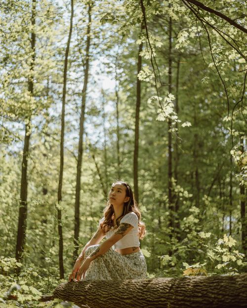 Free Side view of peaceful ethnic female with long wavy hair sitting on trunk in overgrown forest and looking away dreamily Stock Photo