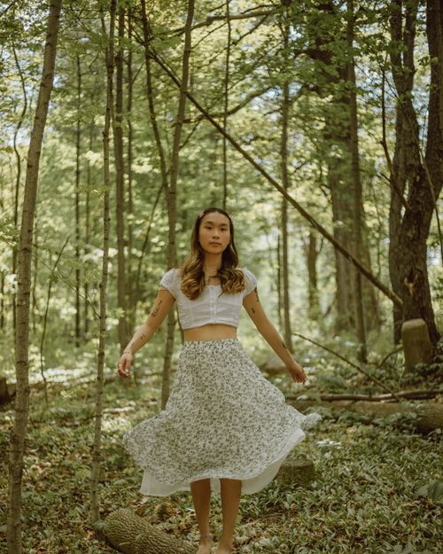 Free Asian female with wavy hair wearing top and flying skirt standing on fallen tree in woodland and looking at camera Stock Photo