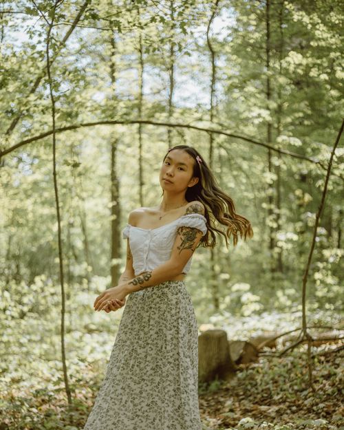Free Tattooed ethnic female with long wavy flowing hair standing among plants in sunny woods and looking at camera Stock Photo