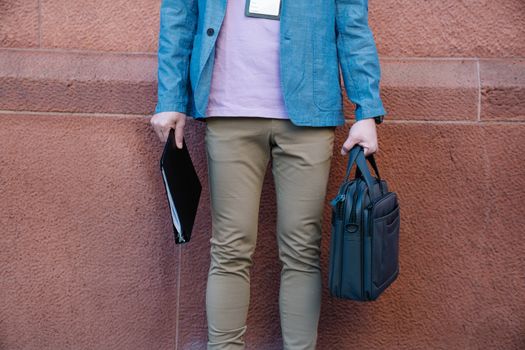 A man standing against a wall holding a briefcase and documents, dressed in casual business attire.