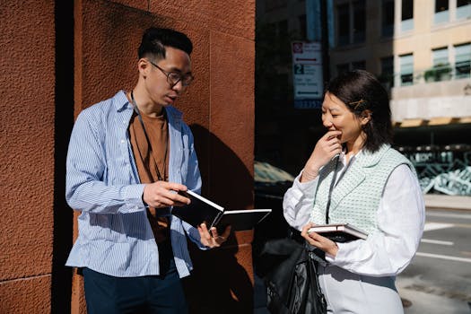 Two young Asian professionals engage in a discussion with notebooks on a city street.