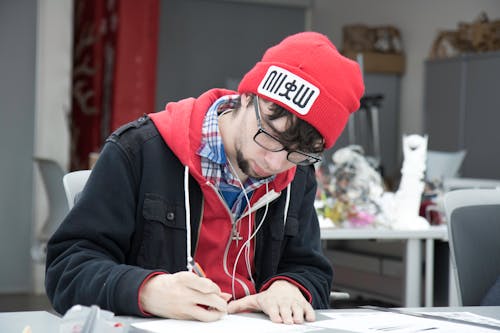 Man in Black Jacket Wearing Red Knit Cap Writing on Paper