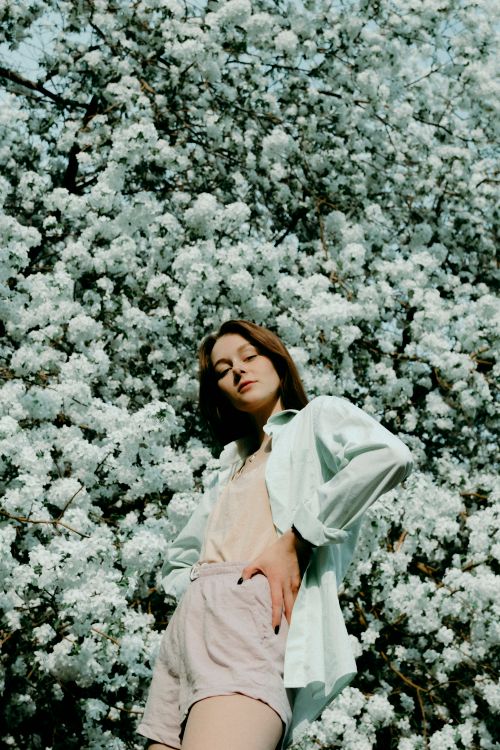 Free From below of slim female in summer outfit standing with hands on waist against blooming tree and looking at camera Stock Photo