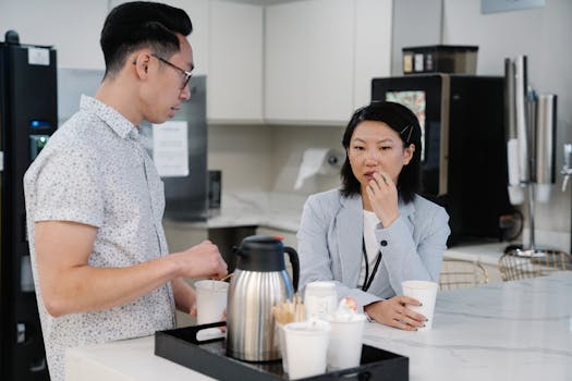 Two colleagues discussing over coffee in a modern office break room setting.