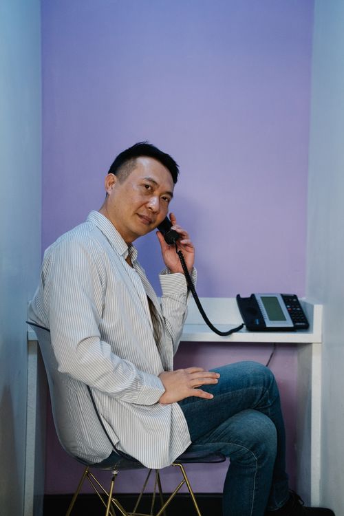 Free Asian man in casual attire casually conversing on an office phone with a purple background. Stock Photo
