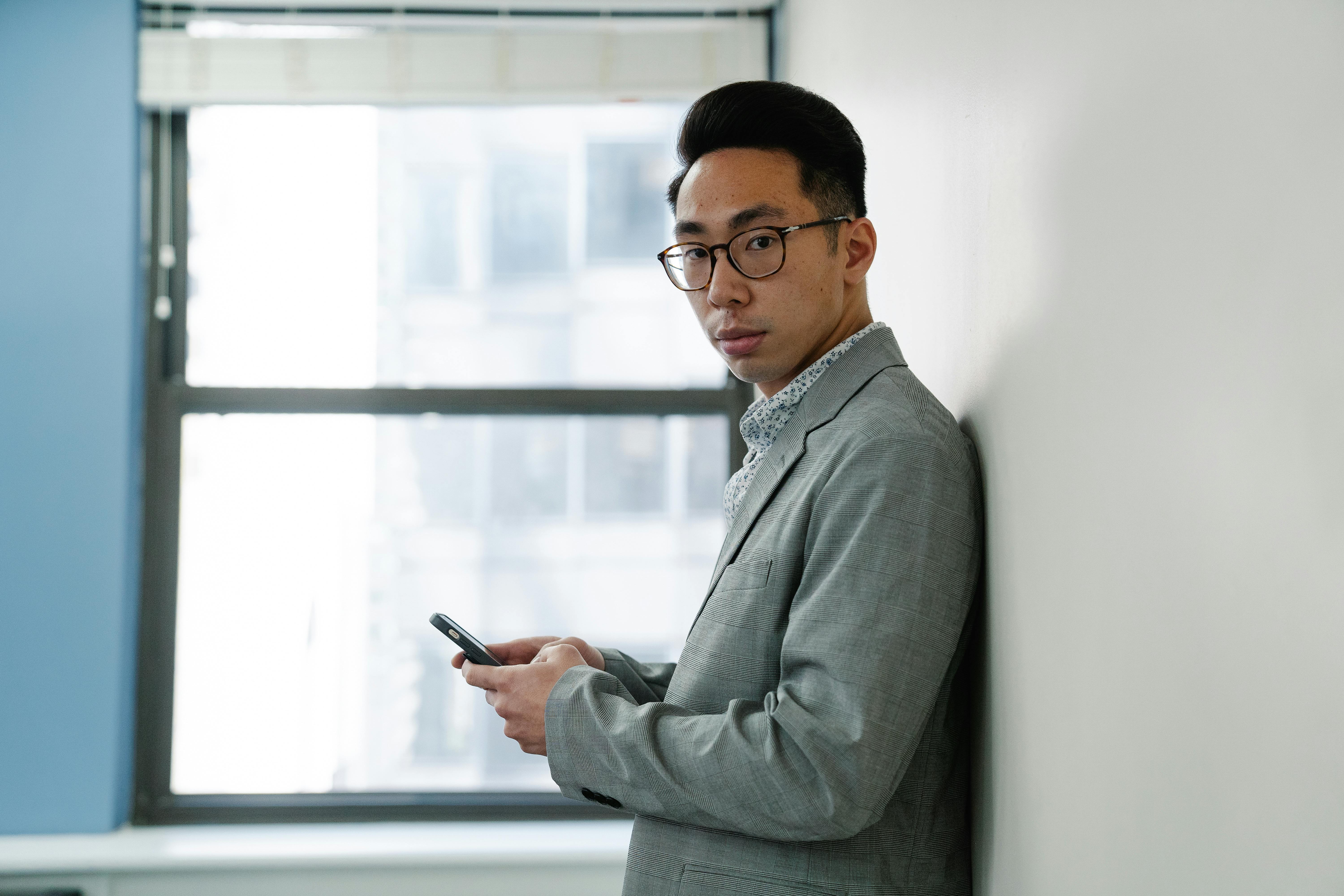 Professionally dressed man using a smartphone by a window, indoors.