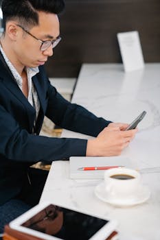 Asian businessman using his smartphone at a cafe table with coffee and tablet, wearing eyeglasses.