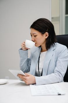 Professional woman in a suit using a digital tablet while enjoying a cup of coffee in an office setting.
