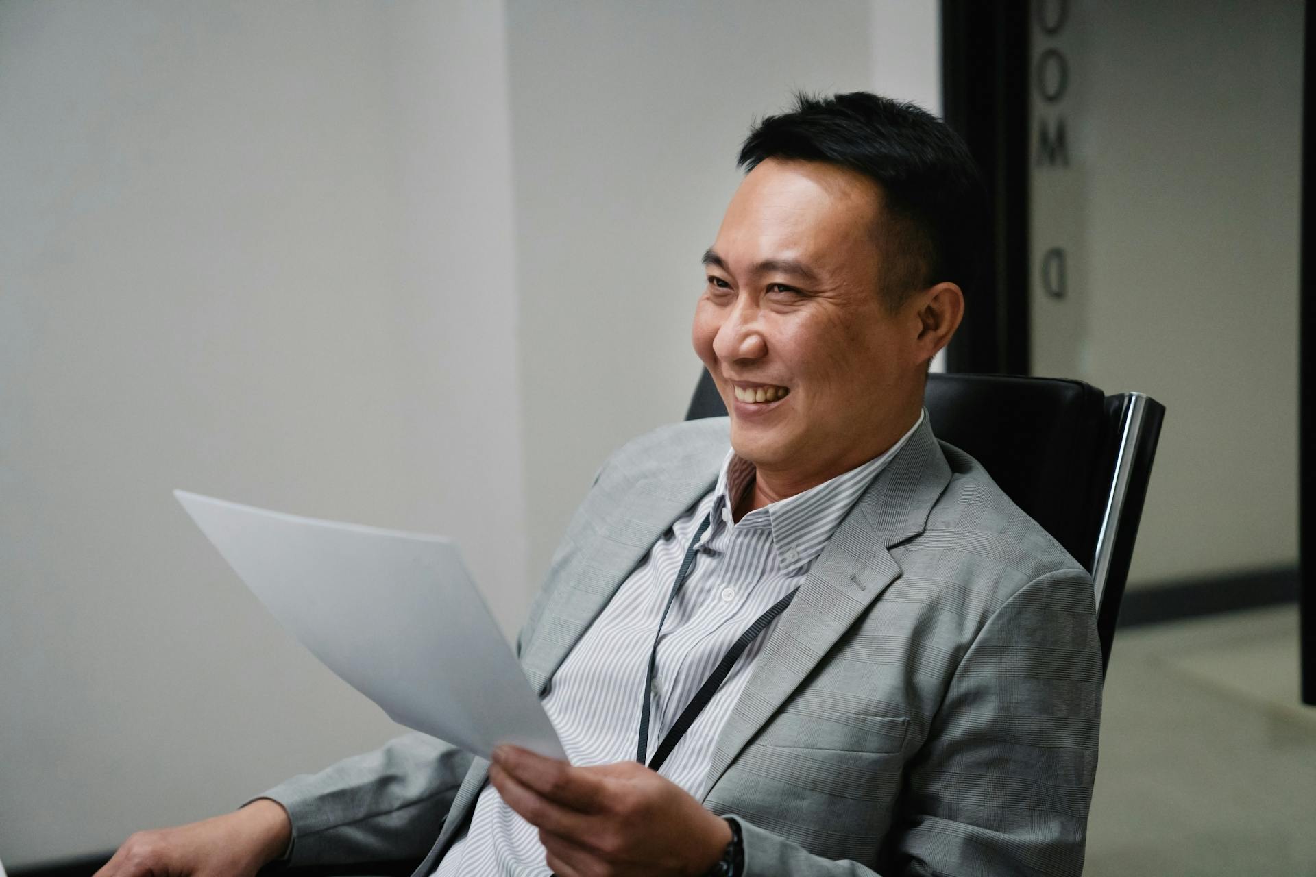 Smiling Asian businessman in grey suit reviews paper documents in modern office setting.