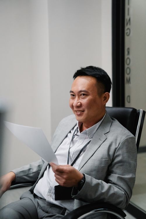 Free Asian man in a gray blazer holding a document while sitting on an office chair indoors. Stock Photo