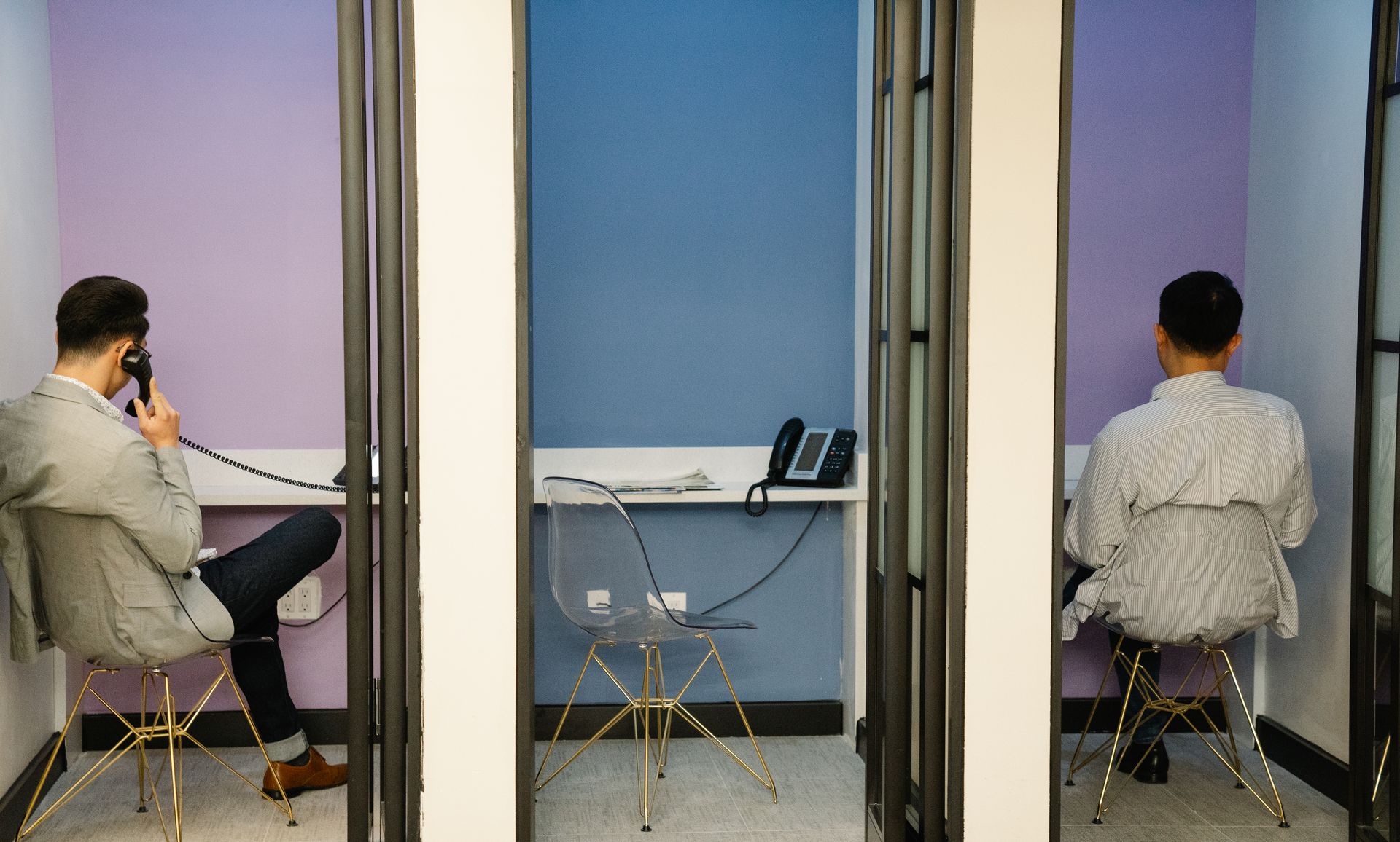 Two businessmen sit in contemporary phone booths using telephones in an office setting.