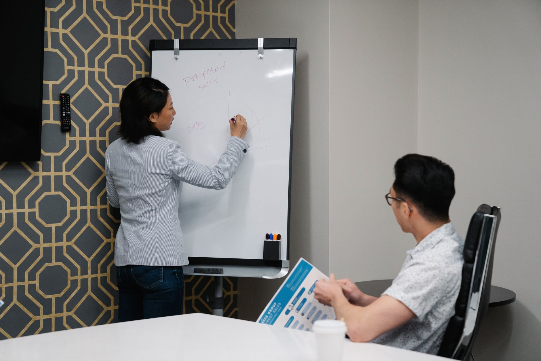 Two colleagues engaged in a business discussion with notes on a whiteboard during a meeting.