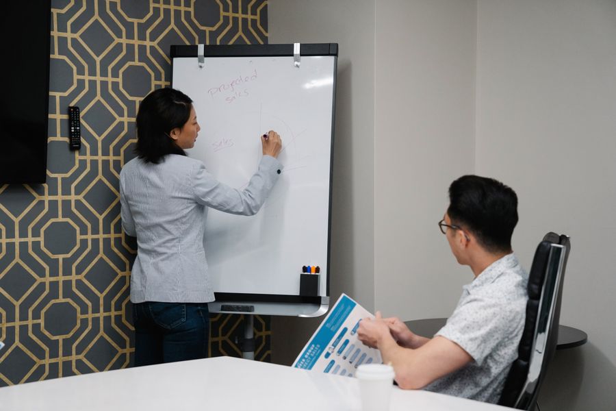 Two colleagues engaged in a business discussion with notes on a whiteboard during a meeting.
