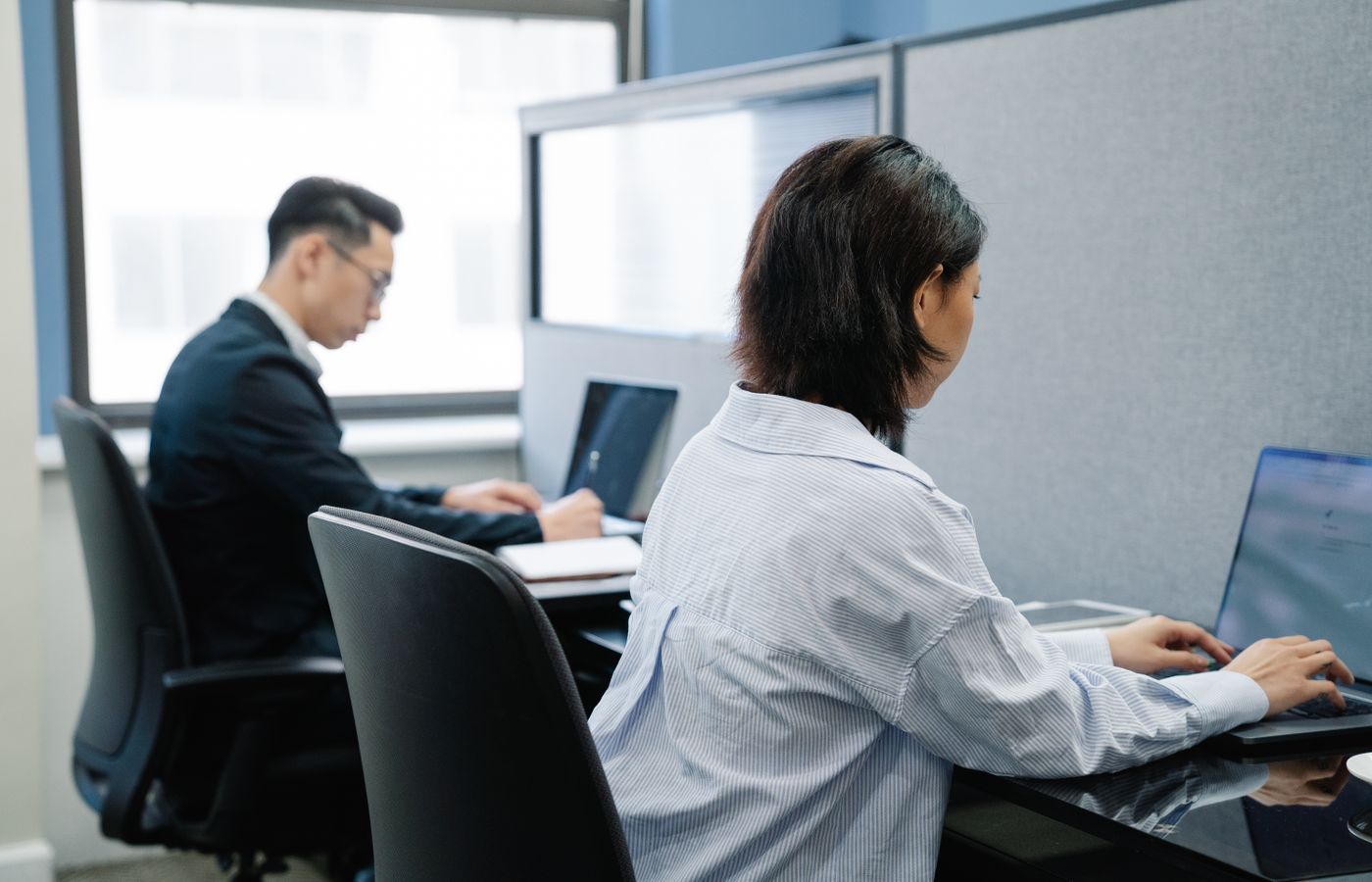 Two professionals working on laptops in a modern office cubicle setup.