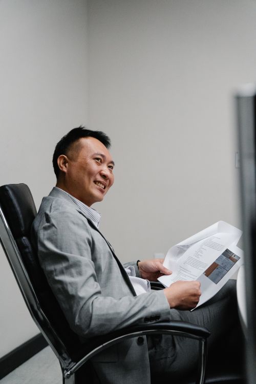 Free Happy man in office attire holding papers and smiling while sitting in a chair indoors. Stock Photo