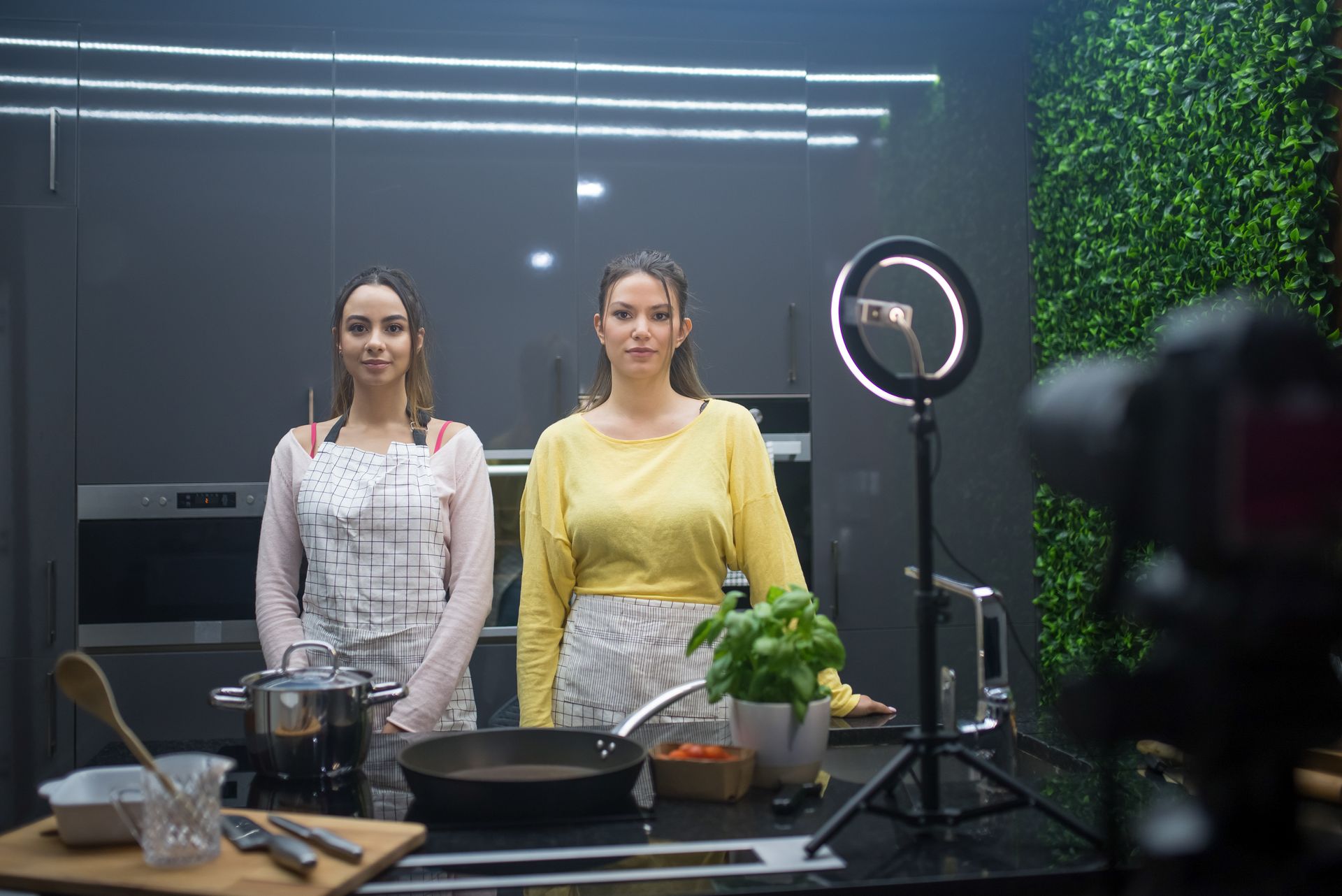 Two Young Women Standing behind the Counter with Cooking Appliances in front of a Camera