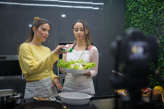 Two women preparing a salad while recording a cooking video in a stylish kitchen setting.