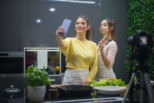 Two women enjoy cooking together in a modern kitchen while capturing the moment with a selfie.