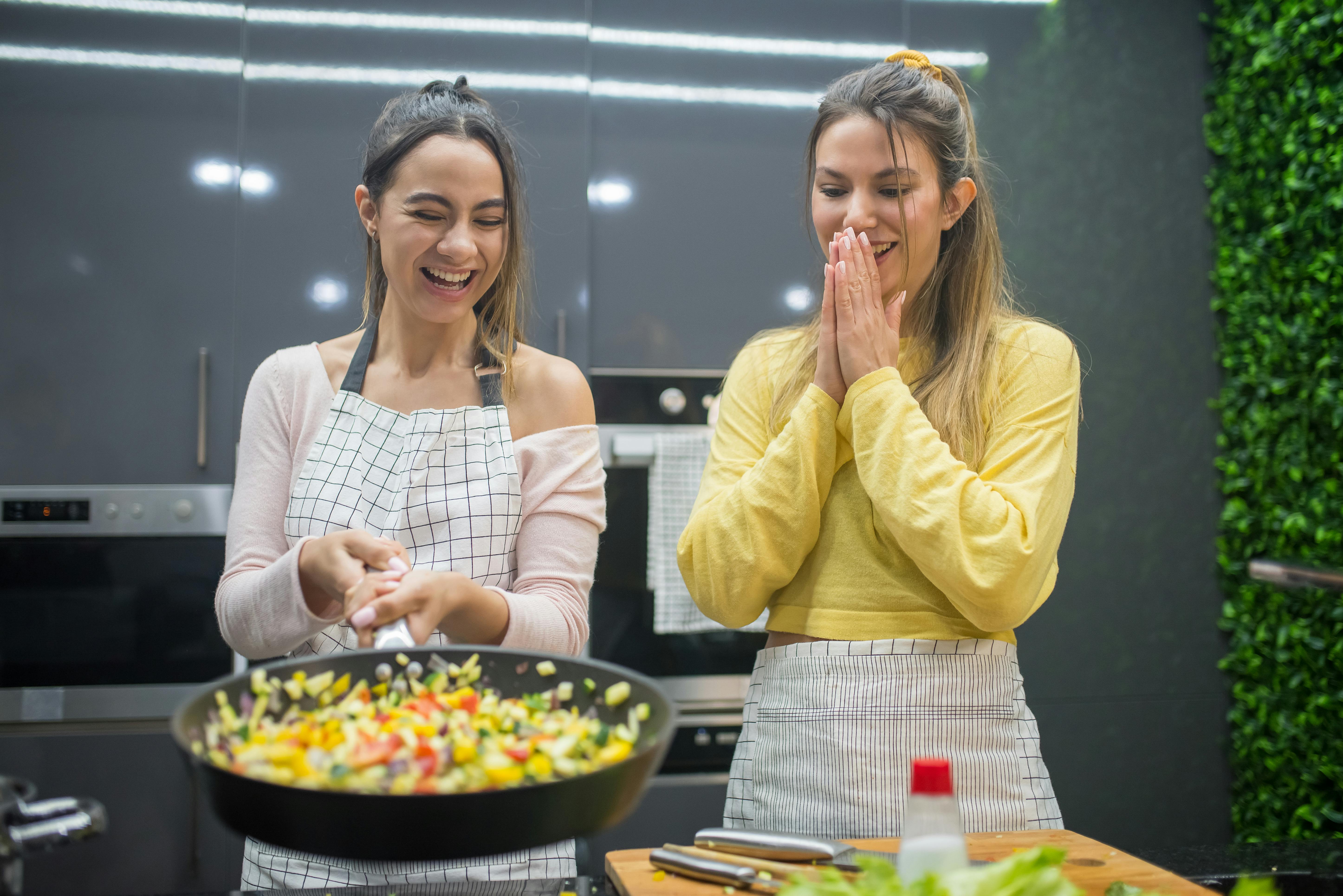Two women enjoying cooking together in a stylish kitchen, laughing and preparing vegetables in a frying pan.