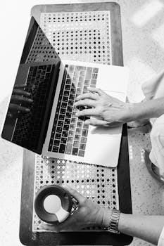 Black and white overhead shot of hands typing on a laptop with a coffee tumbler nearby.