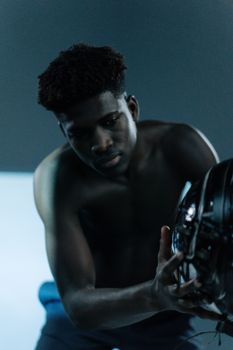 Shirtless young man gazing at a black helmet in a dimly lit indoor setting.